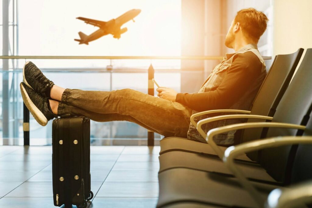 A man sitting at an airport gate waiting for his flight while resting his legs on his suitcase.