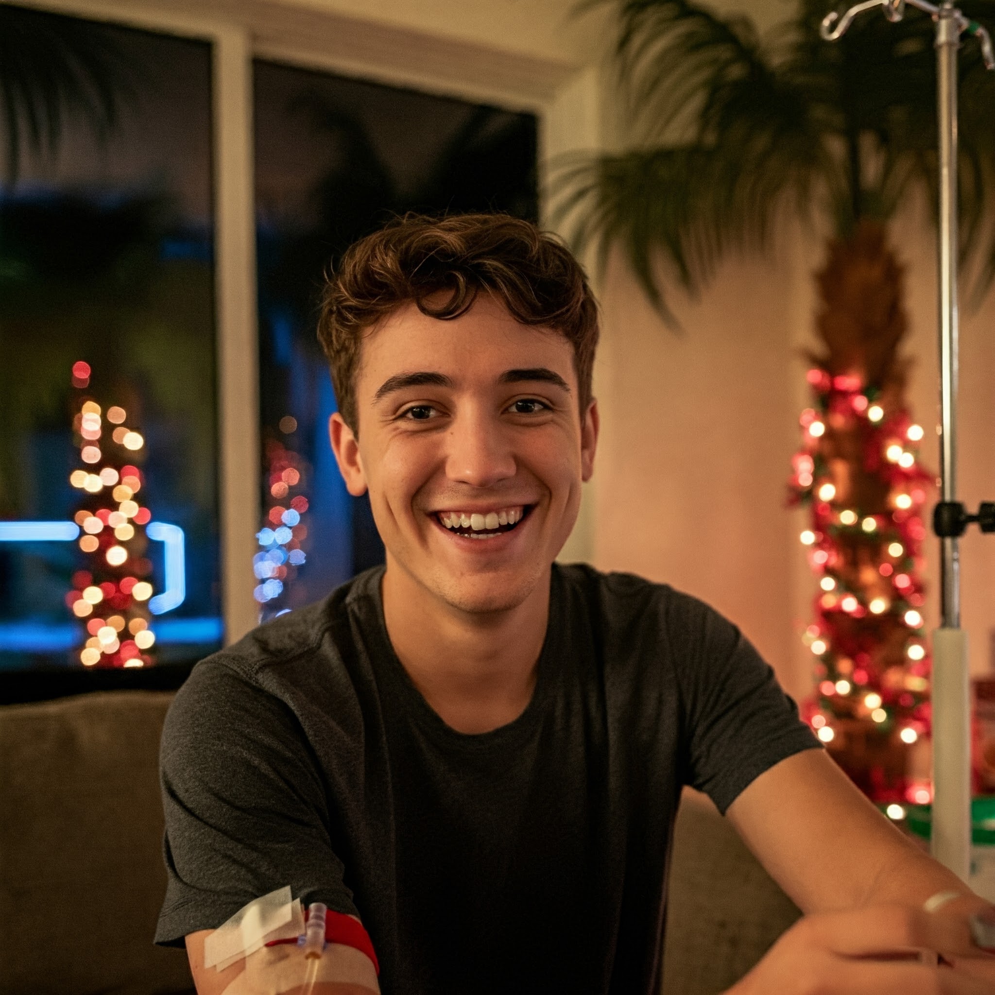 A young man smiles while receiving an IV drip in his festive living room.